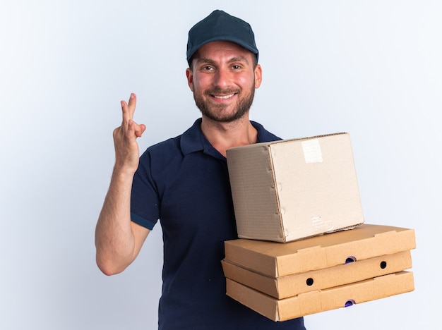 Sorridente giovane fattorino caucasico in uniforme blu e berretto che guarda la telecamera che tiene in mano una scatola di cartone e pacchetti di pizza che ne mostrano tre con la mano isolata sul muro bianco