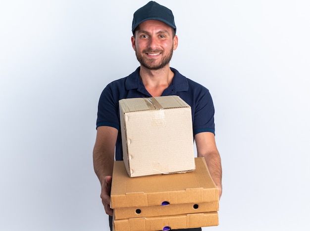 Sorridente giovane fattorino caucasico in uniforme blu e berretto che stende pacchetti di pizza e scatola di cartone verso la telecamera