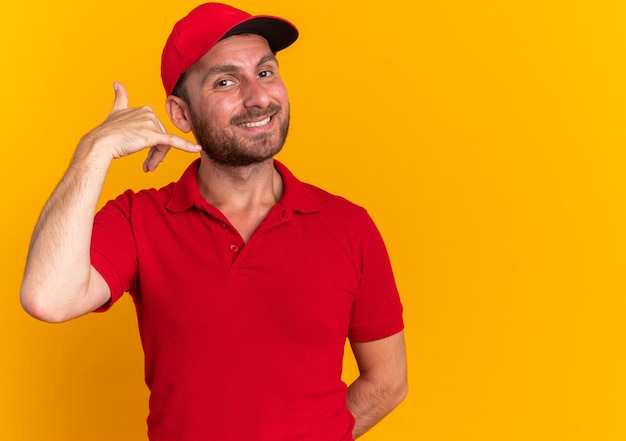 Sorridente giovane fattorino caucasico in uniforme rossa e cappello che tiene la mano dietro la schiena facendo un gesto di chiamata