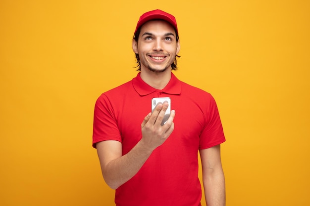 sorridente giovane fattorino che indossa uniforme e cappuccio tenendo il telefono cellulare guardando in alto isolato su sfondo giallo