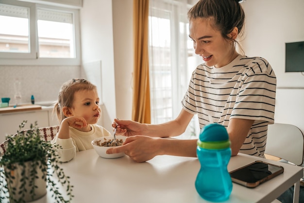 Sorridente giovane genitore femminile che dà un cucchiaio di porridge al suo bambino seduto al tavolo della cucina