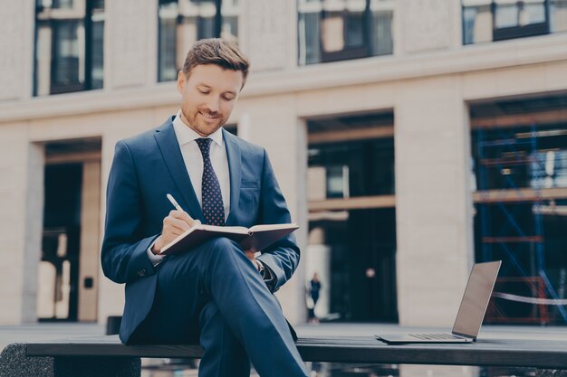 Sorridente giovane imprenditore maschio che lavora al computer portatile all'esterno, prendendo appunti sul taccuino mentre è seduto sulla panchina di fronte all'edificio degli uffici con le gambe incrociate, utilizzando il computer portatile per il lavoro a distanza