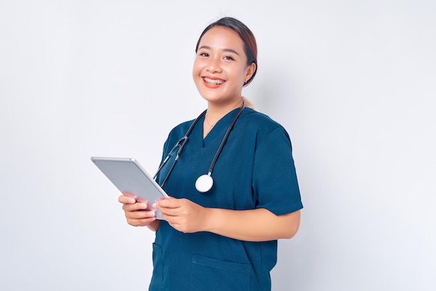 Sorridente giovane infermiera asiatica che indossa un'uniforme blu con uno stetoscopio che tiene una tavoletta digitale isolata su sfondo bianco Concetto di medicina sanitaria