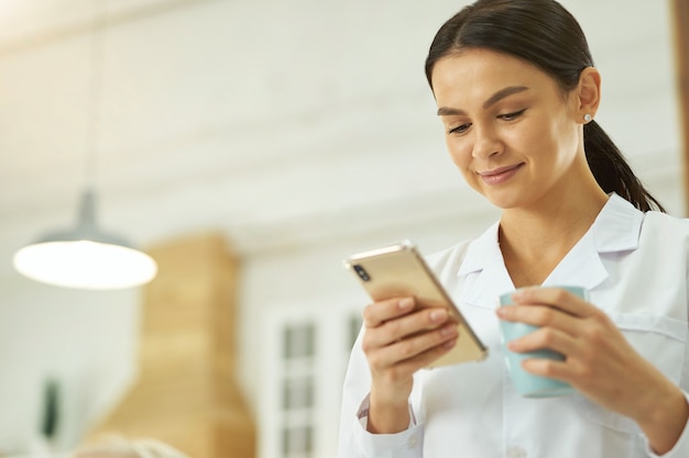 Sorridente giovane infermiera che tiene la tazza e guarda lo schermo del telefono cellulare mentre si trova nella stanza