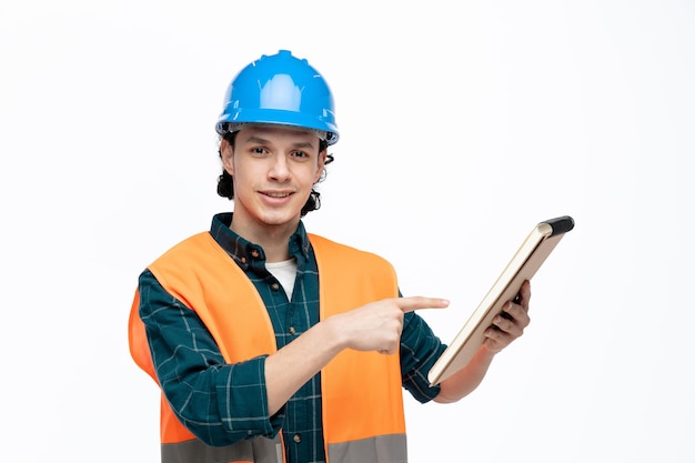 Sorridente giovane ingegnere maschio che indossa casco di sicurezza e giubbotto di sicurezza che tiene e indica il blocco note guardando la fotocamera isolata su sfondo bianco