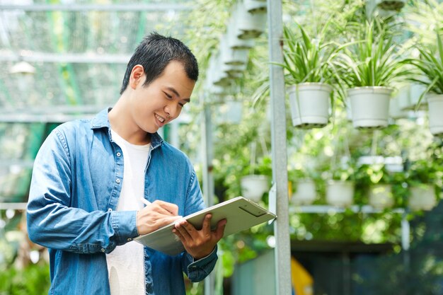 Sorridente giovane lavoratore del centro di giardinaggio che controlla l'elenco dei fiori che sono stati consegnati per la vendita