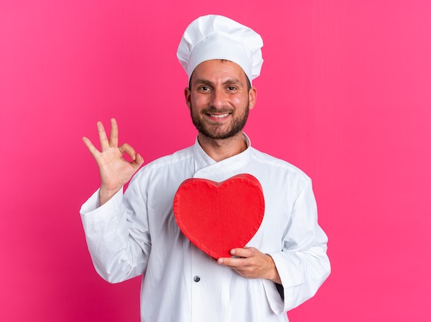 Sorridente giovane maschio caucasico cuoco in uniforme da chef e berretto che tiene a forma di cuore guardando la telecamera facendo segno ok isolato su parete rosa