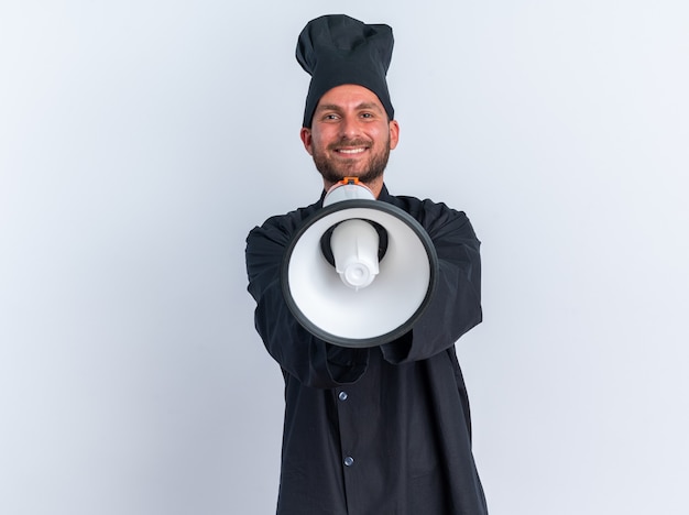 Sorridente giovane maschio caucasico cuoco in uniforme da chef e berretto guardando la telecamera che allunga l'altoparlante verso la telecamera isolata sul muro bianco con spazio di copia