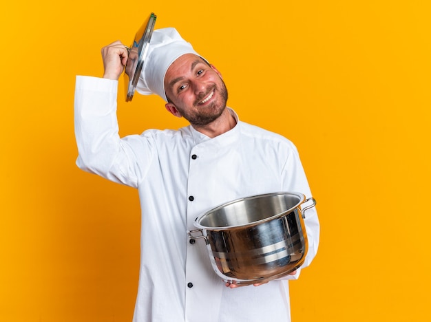 Sorridente giovane maschio caucasico cuoco in uniforme da chef e cappello che tiene pentola e toccando il cappuccio con il coperchio della pentola guardando la telecamera isolata sulla parete arancione