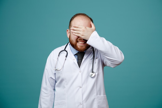 Sorridente giovane medico maschio che indossa cappotto medico e stetoscopio intorno al collo chiudendo gli occhi con la mano isolata su sfondo blu