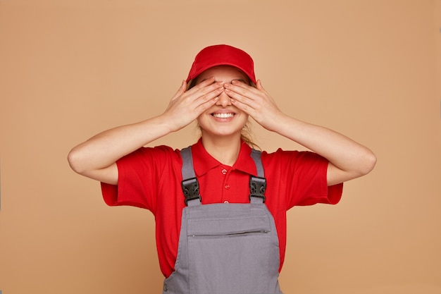 Sorridente giovane operaio edile femminile che indossa uniformi e berretto che copre gli occhi con le mani