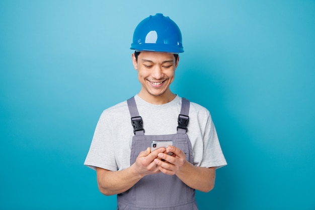 Sorridente giovane operaio edile indossando il casco di sicurezza e tenuta uniforme e guardando il telefono cellulare