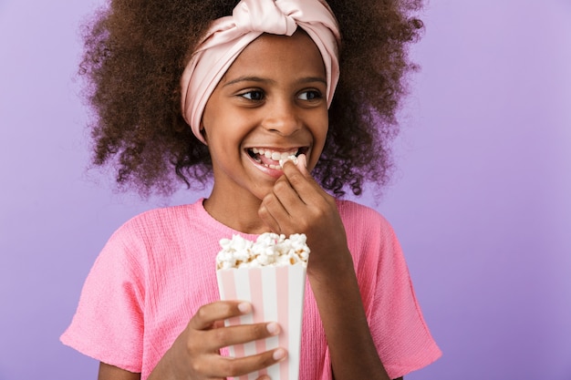sorridente giovane ragazza africana in posa isolata sul muro viola, mangia popcorn.