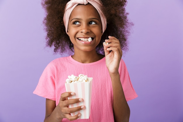 sorridente giovane ragazza africana in posa isolata sul muro viola, mangia popcorn.