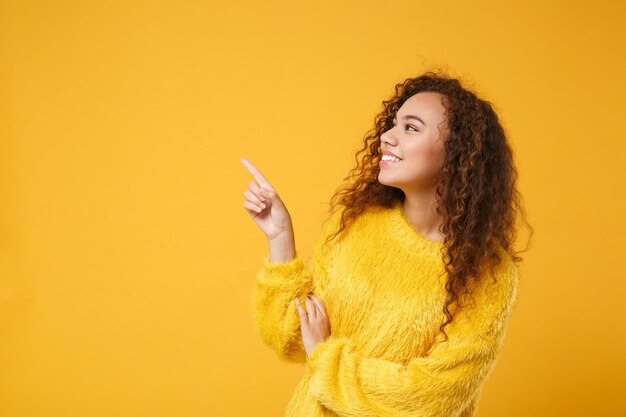 Sorridente giovane ragazza afroamericana in maglione di pelliccia in posa isolata su sfondo giallo arancione in studio. Persone sincere emozioni concetto di stile di vita. Mock up spazio di copia. Puntando il dito indice da parte.