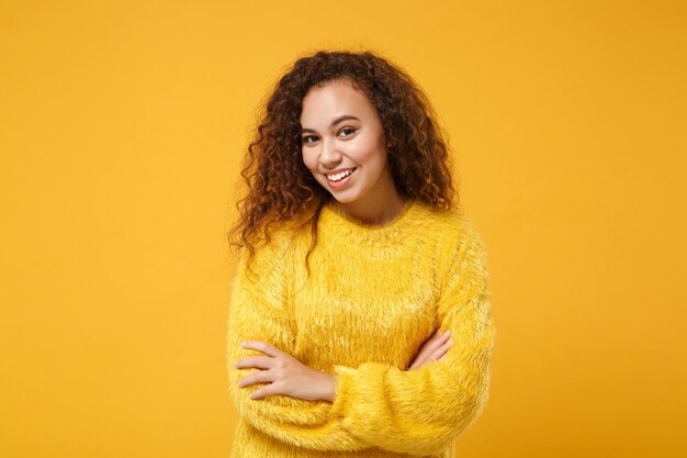 Sorridente giovane ragazza afroamericana in maglione di pelliccia in posa isolata su sfondo giallo arancione, ritratto in studio. Persone sincere emozioni concetto di stile di vita. Mock up spazio di copia. Tenendosi per mano incrociata.