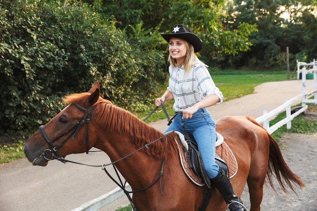 Sorridente giovane ragazza bionda attraente che monta un cavallo all'iarda del cavallo