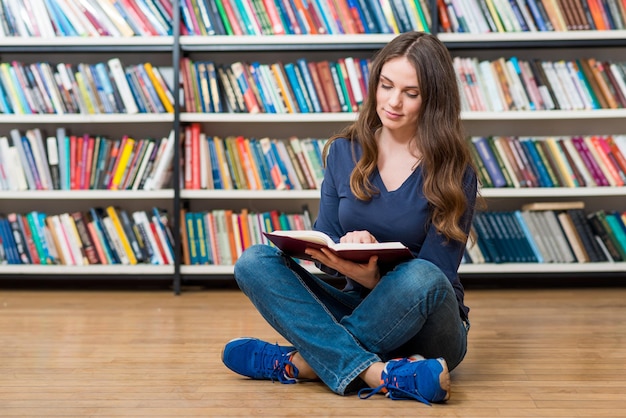 Sorridente giovane ragazza seduta sul pavimento in biblioteca a leggere un libro aperto sulle ginocchia a sinistra con scaffali sfocati sullo sfondo un concetto di lettura