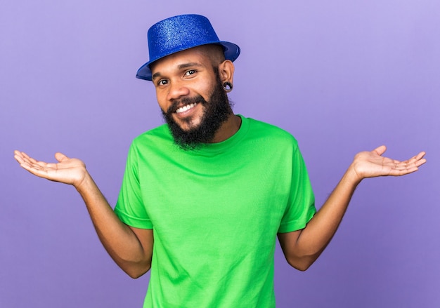 Sorridente giovane ragazzo afroamericano che indossa un cappello da festa allargando le mani isolate sul muro blu