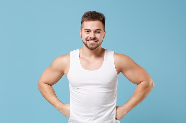 Sorridente giovane ragazzo barbuto 20s in canottiera bianca in posa isolata su sfondo blu pastello parete studio. Concetto di stile di vita sano fitness sportivo. Mock up spazio di copia. In piedi con le braccia sui fianchi in vita.