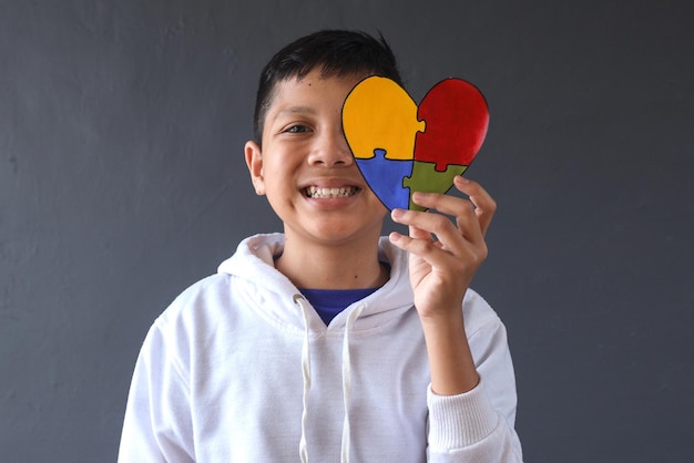 Sorridente giovane ragazzo che tiene a forma di cuore colorato puzzle che copre metà faccia. Giornata Mondiale della Consapevolezza sull'Autismo