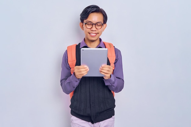 Sorridente giovane studente maschio asiatico di un college in abiti casual con uno zaino utilizzando una tavoletta digitale isolata su sfondo grigio Istruzione nel concetto di college universitario di scuola superiore