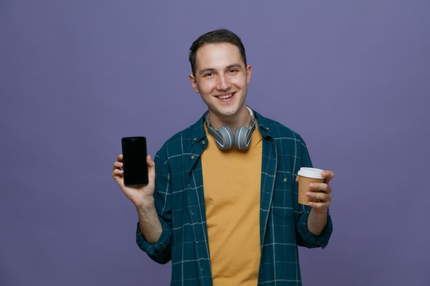 Sorridente giovane studente maschio che indossa le cuffie intorno al collo tenendo una tazza di caffè di carta guardando la fotocamera che mostra il telefono cellulare isolato su sfondo viola