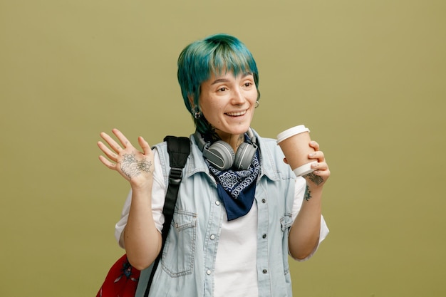 Sorridente giovane studentessa che indossa le cuffie e la bandana sul collo e lo zaino con in mano una tazza di caffè di carta guardando il lato che mostra ciao gesto isolato su sfondo verde oliva