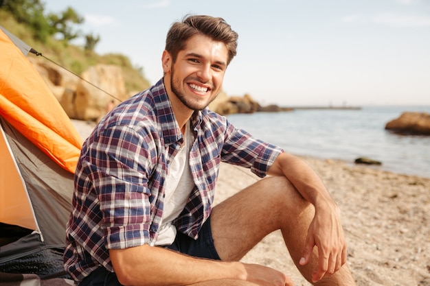 Sorridente giovane turista seduto in tenda turistica in spiaggia e guardando davanti