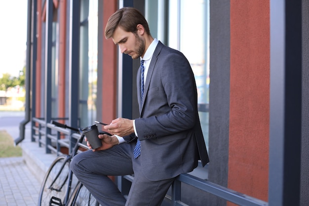 Sorridente giovane uomo alla moda che utilizza il telefono cellulare mentre beve caffè all'aperto con la bicicletta.