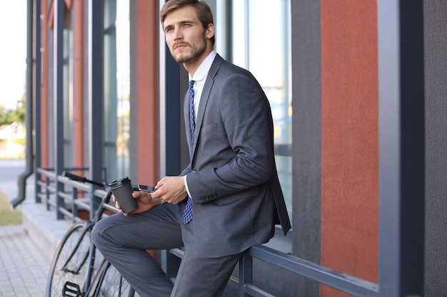 Sorridente giovane uomo alla moda che utilizza il telefono cellulare mentre beve caffè all'aperto con la bicicletta.