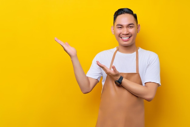 Sorridente giovane uomo asiatico barista barman dipendente che indossa grembiule marrone che lavora nella caffetteria indicando la mano da parte isolata su sfondo giallo Concetto di avvio di piccole imprese