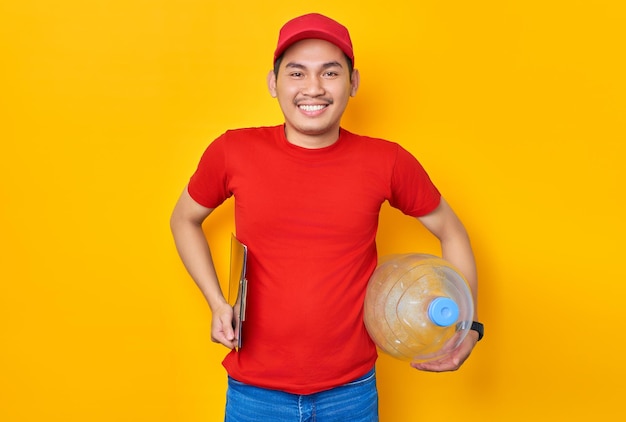 Sorridente giovane uomo asiatico in berretto rosso tshirt uniforme dipendente lavora come rivenditore corriere in possesso di bottiglia d'acqua e appunti con documenti cartacei su sfondo giallo Concetto di servizio di consegna professionale