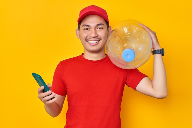 Sorridente giovane uomo asiatico in berretto rosso tshirt uniforme dipendente lavora come rivenditore corriere in possesso di bottiglia d'acqua utilizzando il telefono cellulare isolato su sfondo giallo Concetto di servizio di consegna professionale