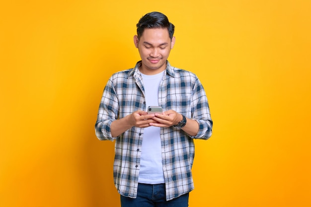 Sorridente giovane uomo asiatico in camicia a quadri utilizzando il telefono cellulare leggendo buone notizie isolato su sfondo giallo