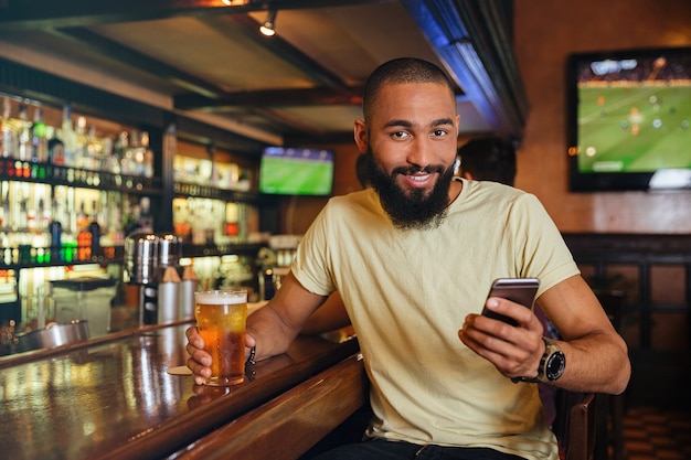 Sorridente giovane uomo che beve birra e usa il cellulare in pub