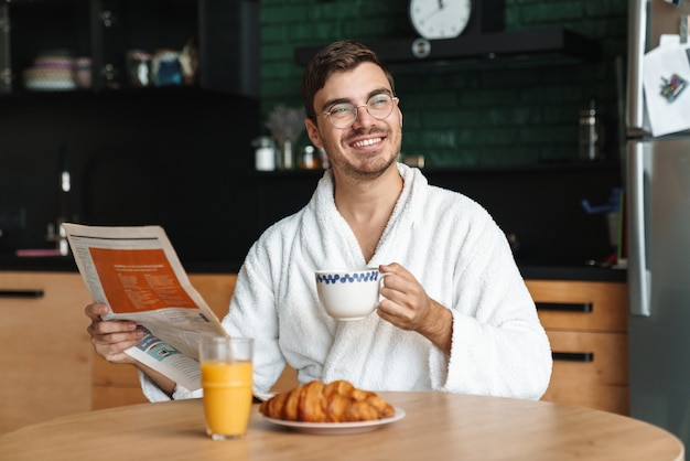 Sorridente giovane uomo che indossa accappatoio fare colazione mentre è seduto in cucina e leggere il giornale al mattino