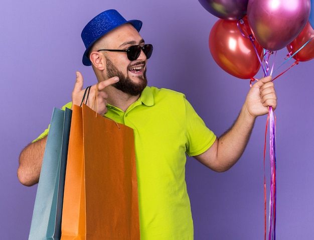 Sorridente giovane uomo che indossa un cappello da festa con occhiali che tengono palloncini con un sacchetto regalo punti a lato