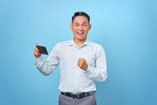 Sorridente giovane uomo d'affari bello che tiene smartphone e guarda la telecamera su sfondo blu