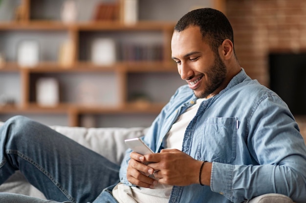 Sorridente giovane uomo di colore utilizzando lo smartphone mentre si riposa sul divano di casa