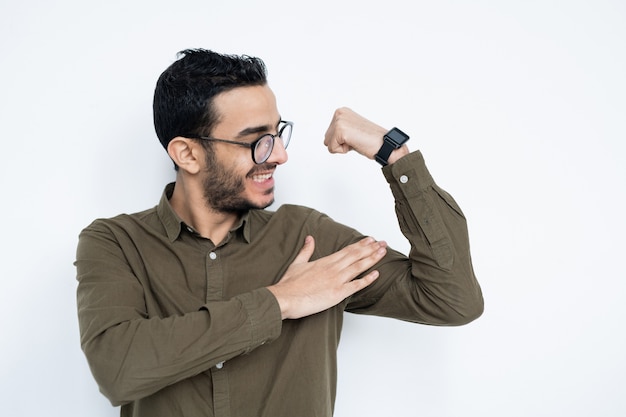 Sorridente giovane uomo forte in occhiali da vista e camicia che tocca i suoi muscoli mentre è in piedi dal muro bianco in studio