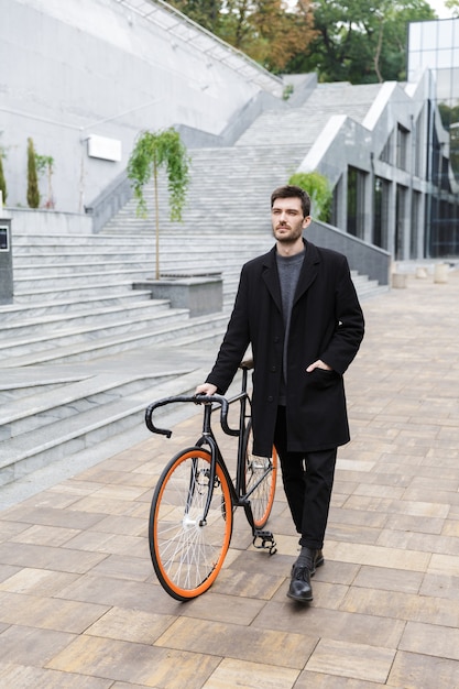 Sorridente giovane uomo vestito in cappotto che cammina con una bicicletta su una strada