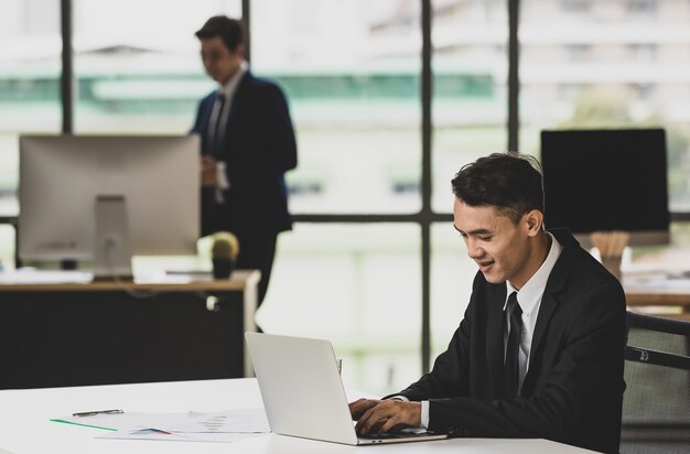 Sorridente imprenditore maschio etnico che digita su netbook e lavora a un progetto sul posto di lavoro con un collega maschio irriconoscibile sfocato