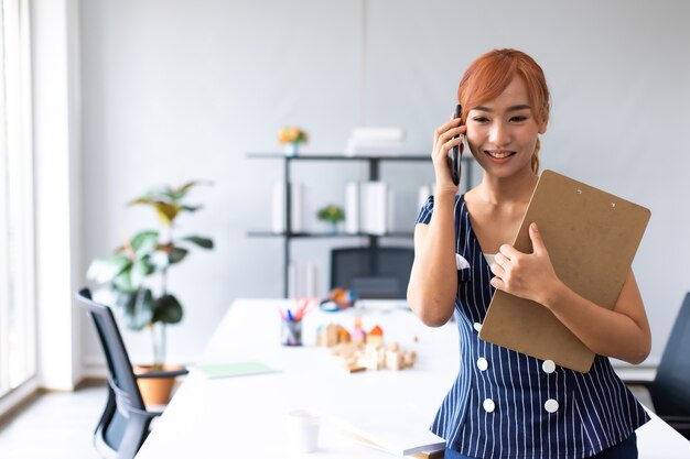 Sorridente imprenditrice asiatica impegnata a lavorare parlando al telefono cellulare in ufficio a casa. Spazio di copia di sfondo banner