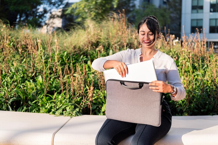Sorridente imprenditrice che prende il suo computer portatile fuori dalla sua valigetta seduta in un parco, concetto di imprenditore digitale e stile di vita urbano, copia spazio per il testo