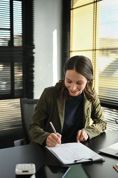 Sorridente investitrice caucasica che firma sul documento aziendale alla sua postazione di lavoro