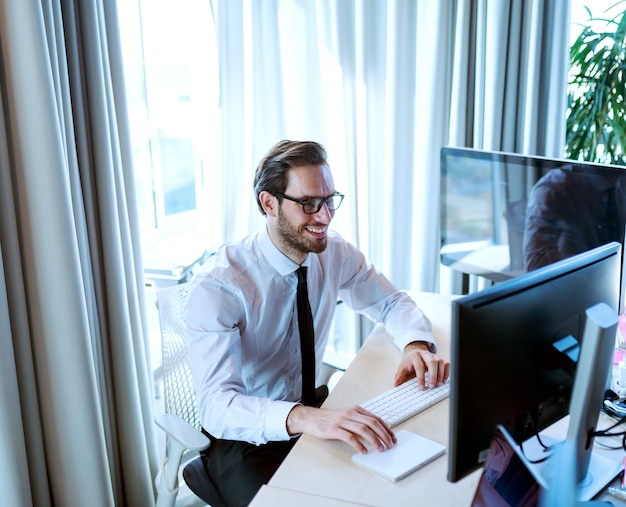 Sorridente lavoratore aziendale in abbigliamento formale utilizzando computer desktop e seduto nel suo ufficio.