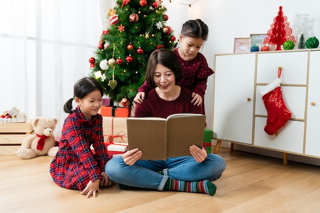 sorridente madre asiatica che legge un libro di fiabe alle sue ragazze in un accogliente soggiorno luminoso con albero di Natale e decorazioni a casa