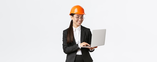 Sorridente manager di fabbrica di ingegnere industriale asiatico femminile di successo in casco di sicurezza e tailleur utilizzando il computer portatile che controlla con il progetto o i progetti sullo schermo