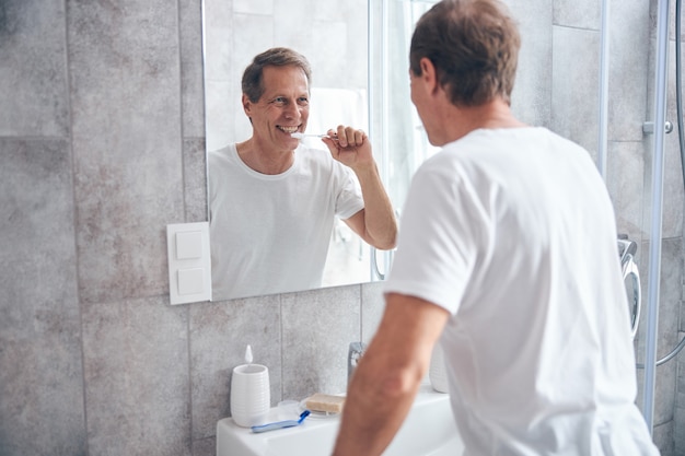 sorridente maschio dai capelli corti che si pulisce i denti davanti allo specchio del bagno